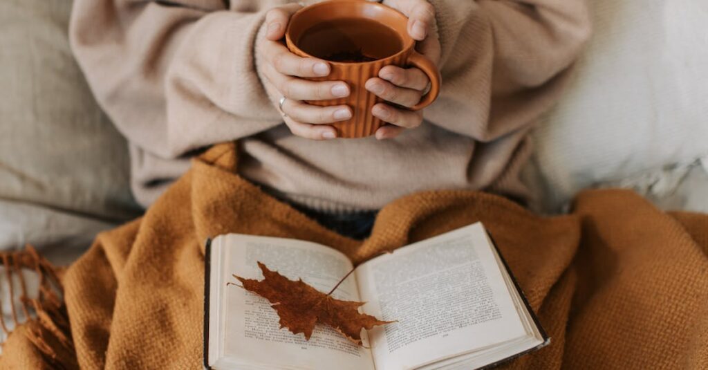 A woman enjoys a cozy autumn day, holding a tea mug over an open book with a maple leaf.