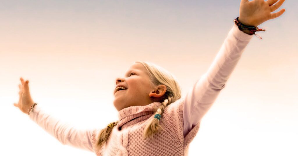 A joyful young girl with braided hair celebrates with arms raised outdoors. Captures happiness and freedom.