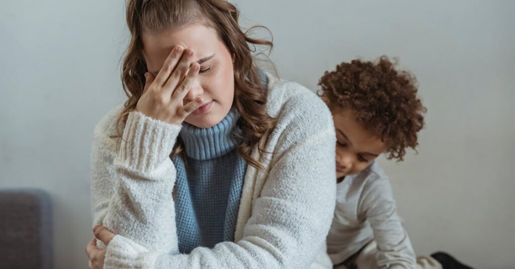 Frustrated mother with hand on forehead and closed eyes sitting near African American son near wall in room at home