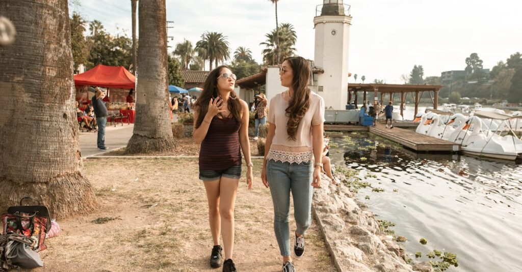 Two young women enjoy a lively conversation while walking by a lakeside lighthouse on a sunny day.